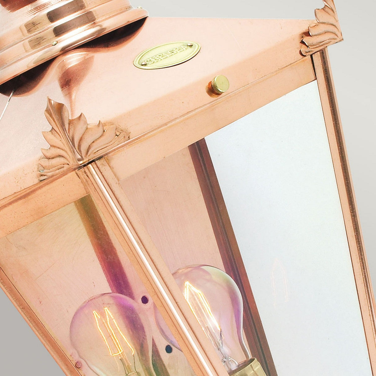 Close-up of a Chelsea Outdoor 1 Light Half Lantern - Copper, featuring decorative leaf details on the corners. Inside, one light bulb enhances its glow, reflecting against the lantern's glass panels. The metallic copper surface has a soft sheen, making it an elegant piece for outdoor lighting against a neutral background.