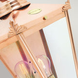 Close-up of a Chelsea Outdoor 1 Light Half Lantern - Copper, featuring decorative leaf details on the corners. Inside, one light bulb enhances its glow, reflecting against the lantern's glass panels. The metallic copper surface has a soft sheen, making it an elegant piece for outdoor lighting against a neutral background.