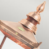 Close-up of the Chelsea Outdoor 1 Light Grande Up Wall Lantern - Copper, featuring a pointed top and glass panels. This copper lantern is adorned with an elegant decorative finial, a brass plaque, and has an IP44 rating. The background is a soft gray.