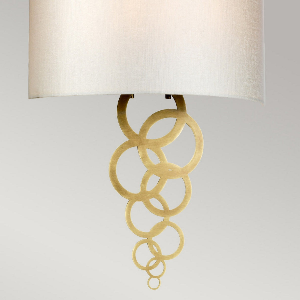 The Curtis Large Wall Light in aged brass is a decorative lighting fixture featuring a white shade and intricate gold metal accents with interlocking circular rings extending downward, all set against a contemporary neutral gray background.