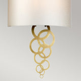 The Curtis Large Wall Light in aged brass is a decorative lighting fixture featuring a white shade and intricate gold metal accents with interlocking circular rings extending downward, all set against a contemporary neutral gray background.