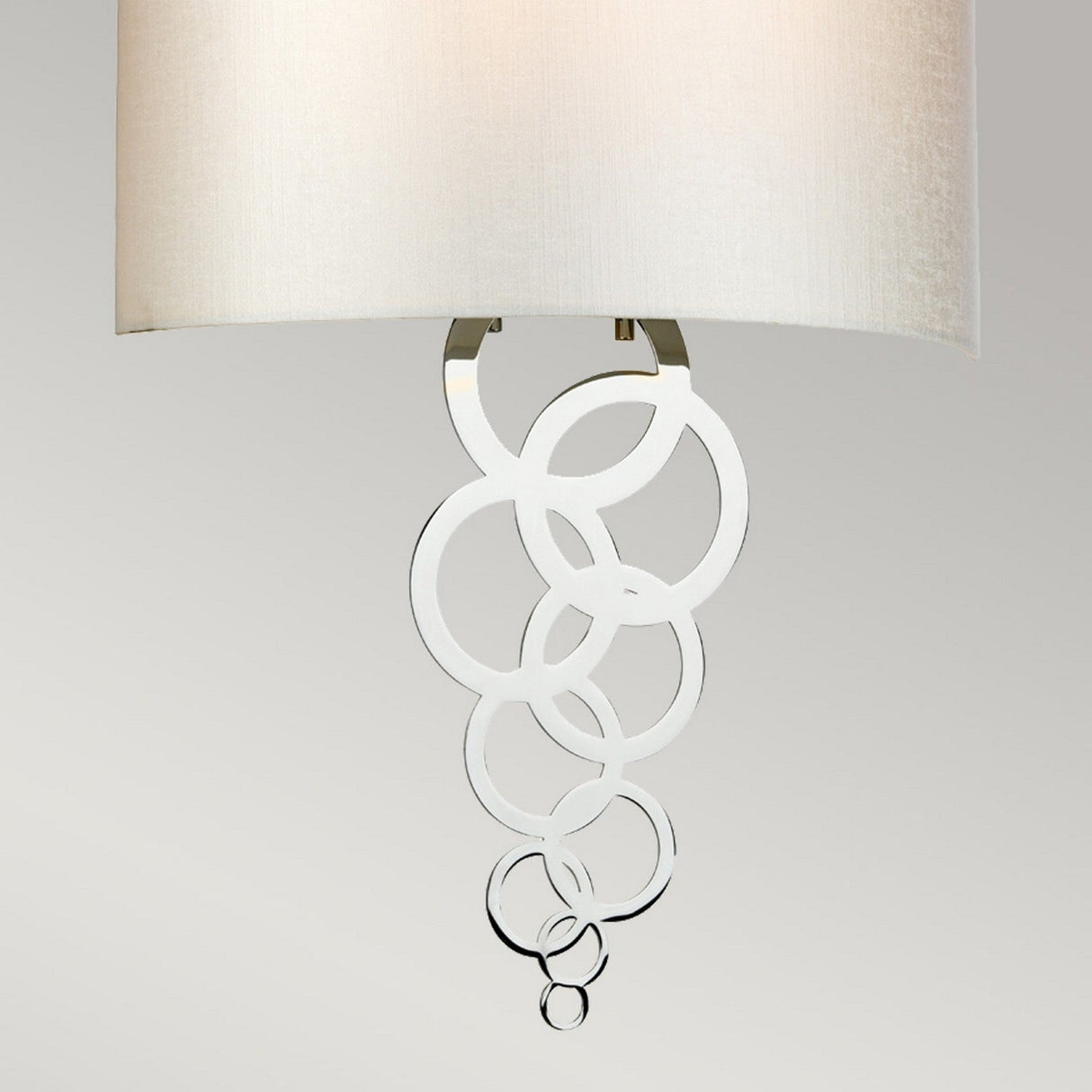 The Curtis Large Wall Light - Polished Chrome is a contemporary fixture featuring a cream-colored shade and a decorative polished chrome accent below, which displays interlocking circles of various sizes against a sleek gray background.