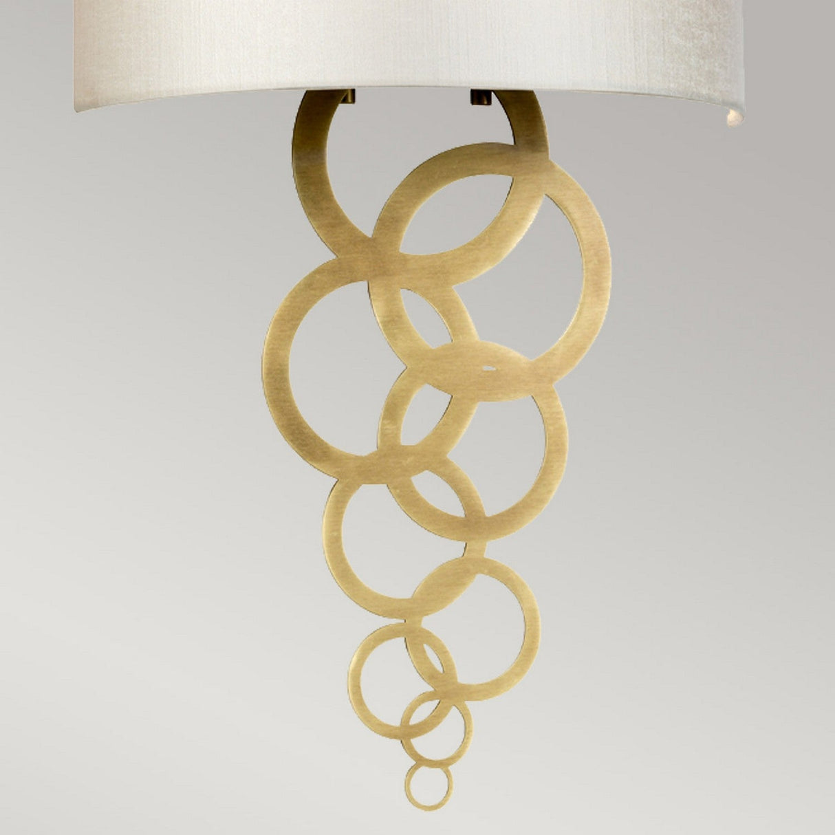 The Curtis Medium Wall Light - Aged Brass showcases modern design with its aged brass rings gracefully overlapping a beige lampshade set against a neutral background.