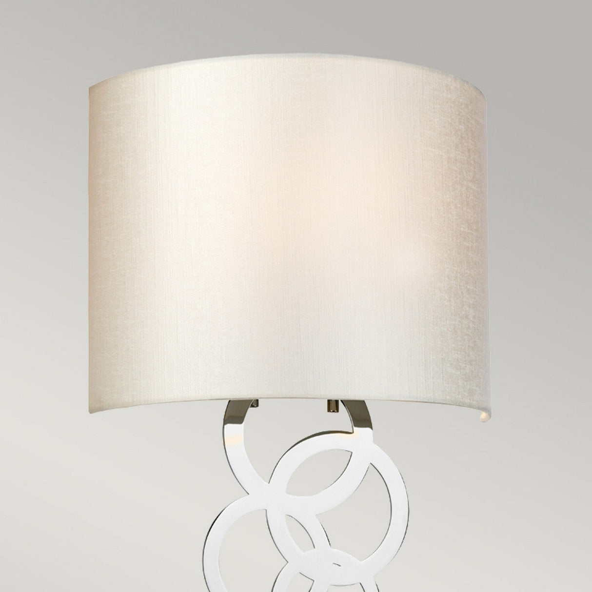 A contemporary desk lamp featuring a spacious cylindrical beige lampshade and a base adorned with interlocking metal circles, this piece captures the essence of modern illumination. Its sleek design enhances any room, much like the refined Curtis Medium Wall Light in polished chrome adds sophistication to an elegant setting.