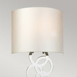 A contemporary desk lamp featuring a spacious cylindrical beige lampshade and a base adorned with interlocking metal circles, this piece captures the essence of modern illumination. Its sleek design enhances any room, much like the refined Curtis Medium Wall Light in polished chrome adds sophistication to an elegant setting.