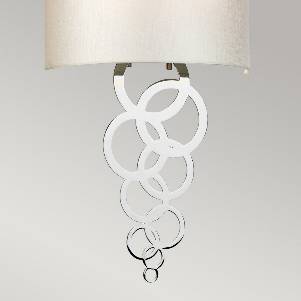 The Curtis Medium Wall Light in polished chrome features a contemporary cascading circular metal design beneath a cream shade. The varying circle sizes contribute to its elegant and sophisticated modern lighting aesthetic.