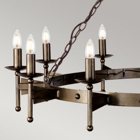 The Cromwell 12 Light Chandelier - Old Bronze offers a rustic elegance with its circular, handcrafted solid iron frame and twelve candle-shaped light bulbs. Exuding medieval charm, it is suspended by a chain that enhances its Old Bronze finish against a simple gray background.