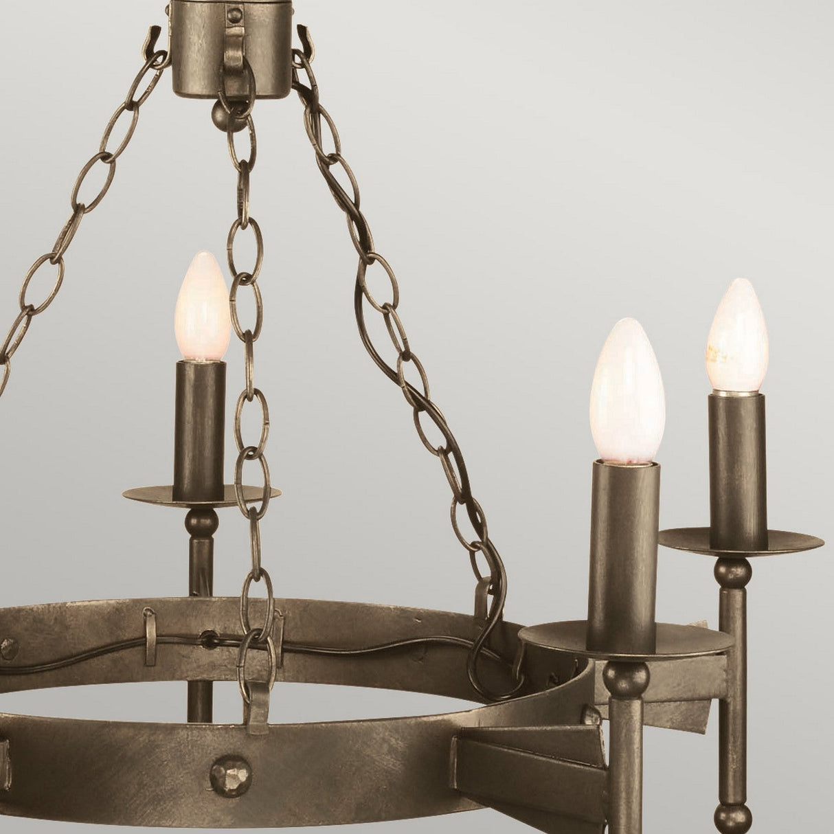 Close-up of a Cromwell 5 Light Chandelier - Old Bronze with a medieval-inspired design, showcasing a circular frame and five candle-like light bulbs. This handcrafted iron fixture is suspended by a series of metal chains against a neutral light gray background.