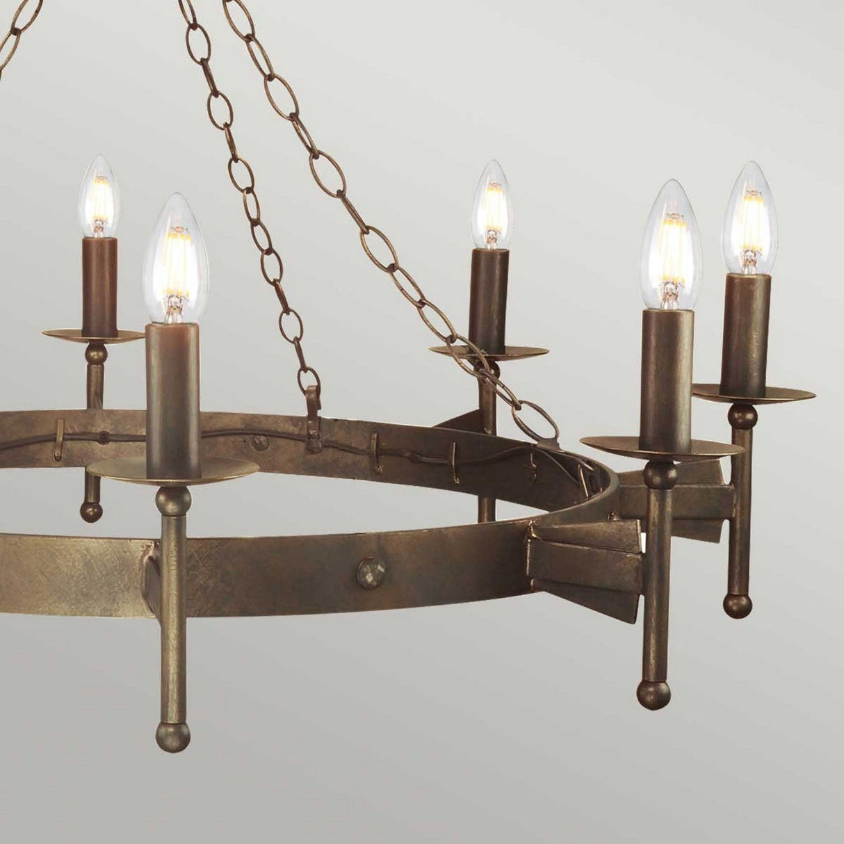 The Cromwell 8 Light Chandelier in an old bronze finish features a medieval lighting design, with chains holding the fixture and showcasing eight exposed light bulbs. This wrought iron piece stands against a plain gray background, adding timeless elegance to any room.