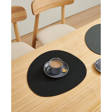 Dahej Placemat in black, a rounded pebble-shaped mat with matt felt-like texture, shown on a wooden table under a grey cup and saucer.
