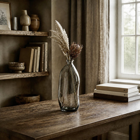 Dahukeng Vase in smoked grey glass with a curved, leaning bottle shape and slim neck, holding dried pampas grass on a wooden desk by a window.