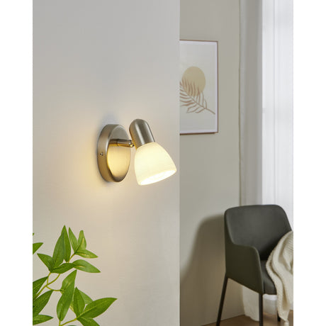 Dakar 1 wall spotlight in satin nickel with a round backplate and jointed arm holding a white glass shade, shown lit on a wall in a living room setting.