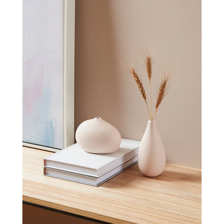 Daraina Ceramic Vase in matt pink, teardrop-shaped with a slender neck holding dried wheat stems, styled on a wooden shelf beside a rounded pink vase and stacked books.
