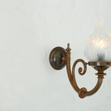 Darwin Glass Wall Light with a brass scrolled arm and round backplate, holding a frosted flame-shaped glass shade lit from within, against a plain light background.