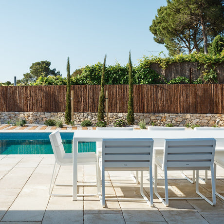 White Dats aluminium outdoor dining table with slim straight legs, paired with matching white dining chairs beside a pool and stone-walled garden.