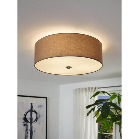 Dernessa 3 Light Ceiling Light with taupe fabric drum shade, satin nickel trim and white glass diffuser, shown glowing in a living room with a plant and curtains nearby.