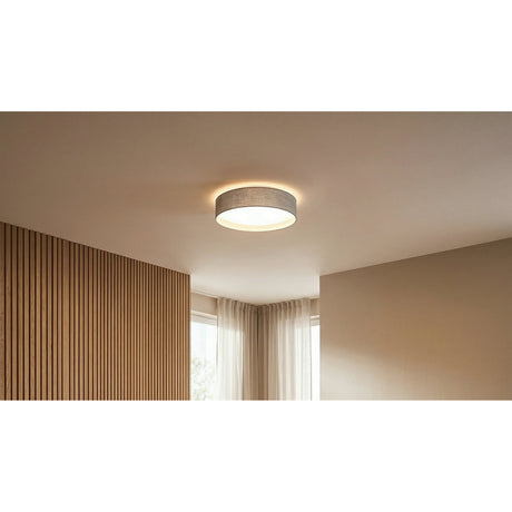 Dernessa LED Flush Ceiling Light in taupe fabric with white diffuser, shown glowing on a room ceiling above wood-panelled walls and sheer curtains.