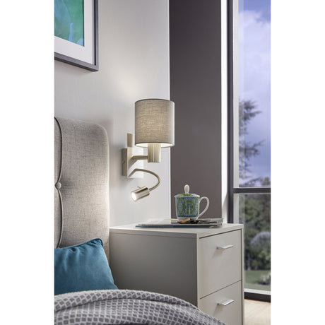 Dernessa LED Wall Light 15cm in satin nickel with a grey fabric drum shade and flexible reading light arm, shown above a bedside table in a bedroom setting.