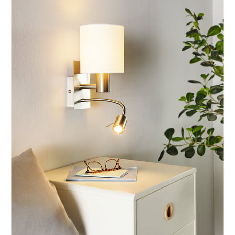 Dernessa LED Wall Light with white fabric cylinder shade and satin nickel body, featuring a flexible reading arm, mounted above a bedside cabinet.
