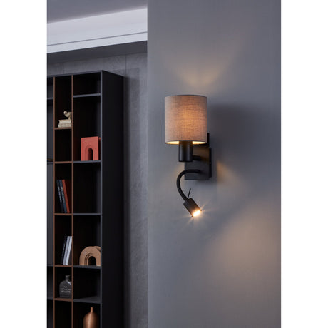 Dernessa LED Wall Light in black steel with an anthracite-brown fabric drum shade and a flexible arm ending in a small LED reading spot, mounted on a grey wall beside a dark bookshelf.