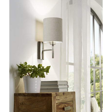 Dernessa Wall Light 14.5cm in satin nickel with a cylindrical grey fabric shade, mounted above a wooden console table near a sheer-curtained window.