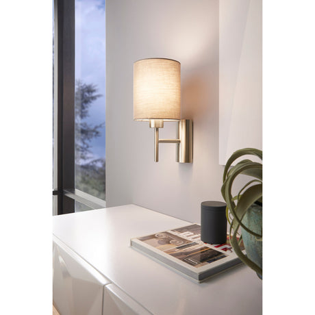 Dernessa Wall Light in satin nickel with a cylindrical taupe fabric shade, mounted above a desk in a living room setting.