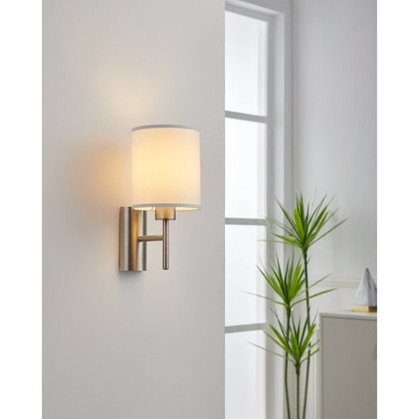 Dernessa Wall Light 14.5cm in satin nickel with a white fabric drum shade, shown mounted beside a window in a living room with a plant nearby.