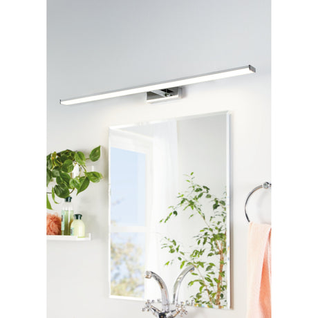 Dervette LED Bathroom Wall Light, a slim 120cm chrome and silver bar fixture with central mount, shown above a mirror in a bathroom setting with a plant and towel ring nearby.