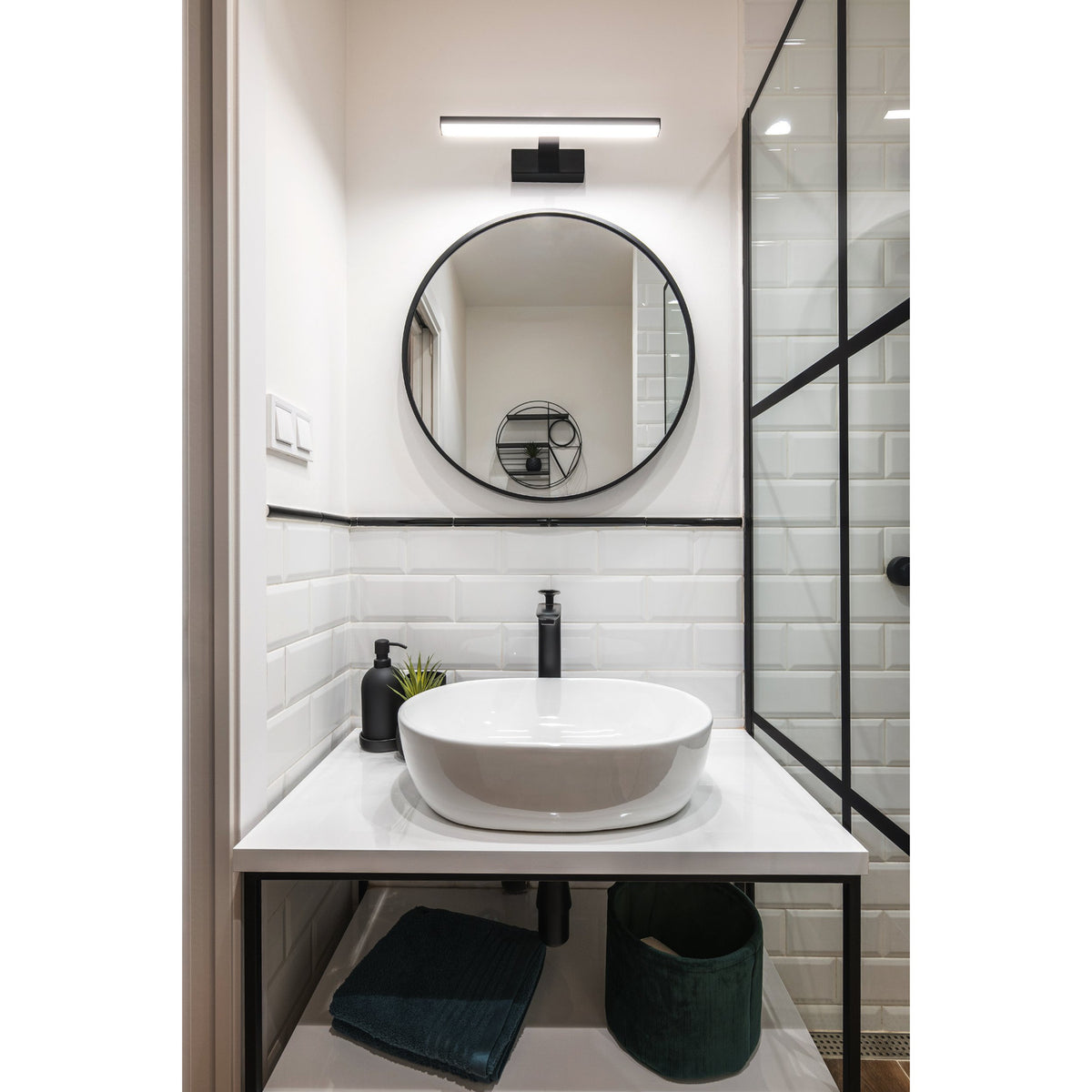 Dervette LED Bathroom Wall Light in black, a slim 40cm bar with white diffuser above a round mirror in a white-tiled bathroom with vessel sink and glass shower screen.