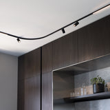 Deva 24V surface-mounted lighting track in black, curving across a ceiling with adjustable spotlights and a linear light, above dark wood cabinetry.