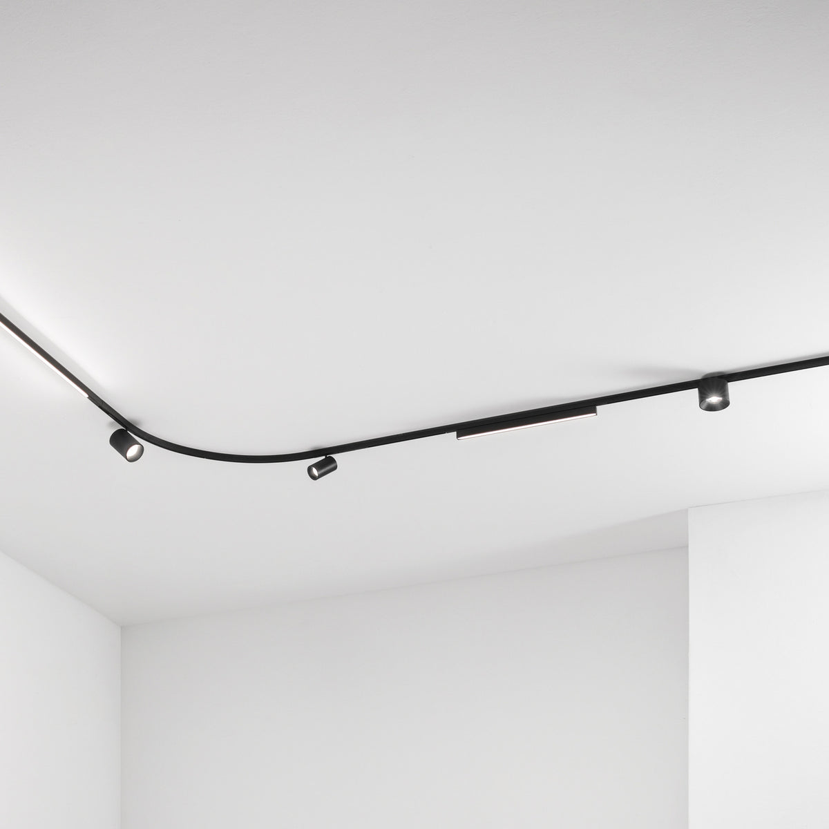 Deva 2m Surface 24V Lighting Track in black, shown curving across a white ceiling fitted with spotlights and a linear light module.
