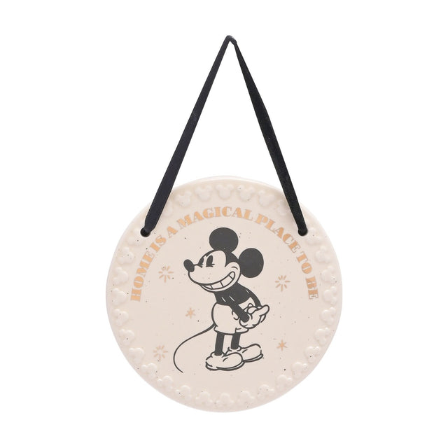 Round white ceramic wall plaque with a black Mickey Mouse illustration, gold "Home is a magical place to be" text, embossed Mickey head border, and black ribbon hanger on a plain background.