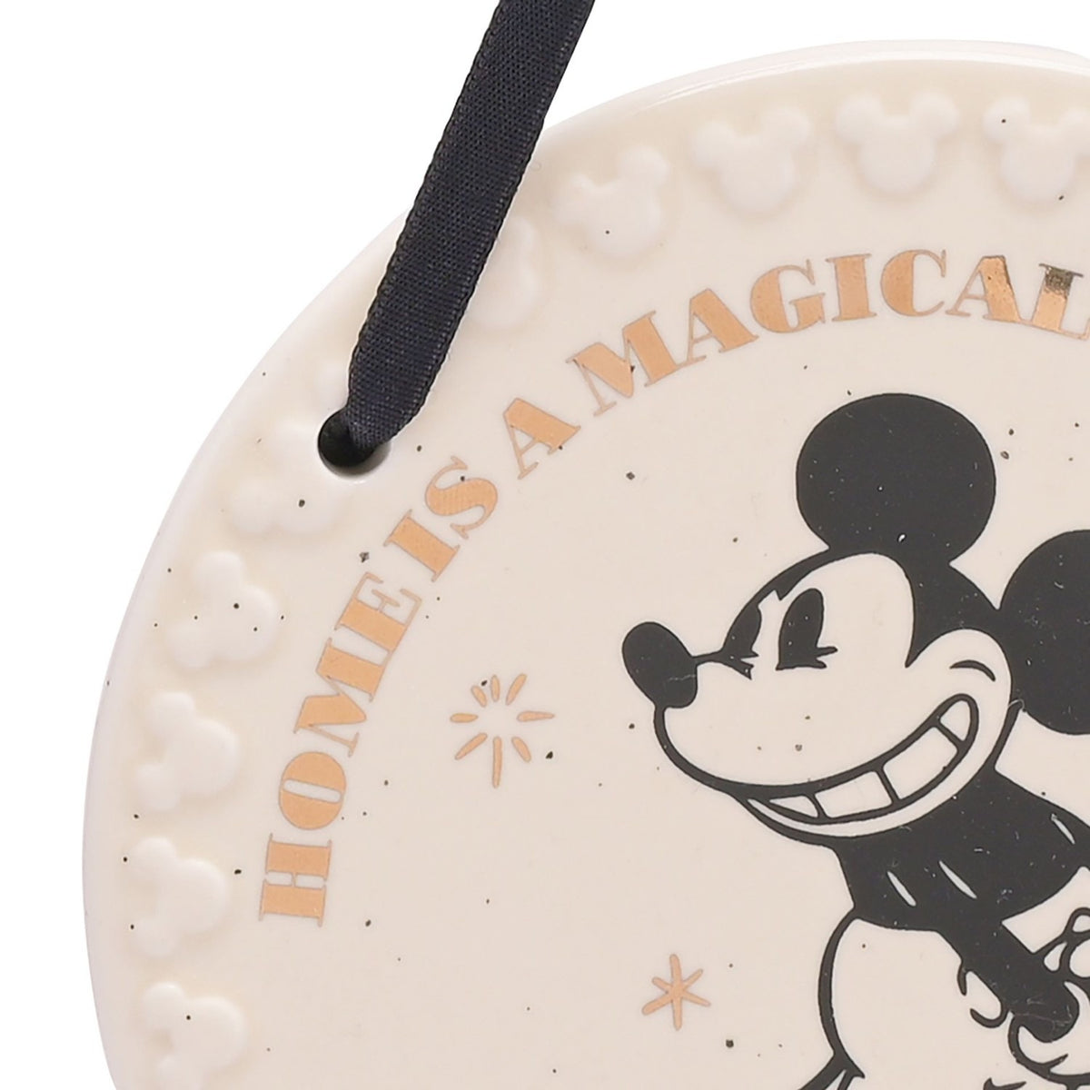 Close-up of round white ceramic Mickey Mouse wall plaque with black ribbon hanger, gold "Home is a Magical..." text, and raised Mickey head border detail.