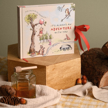 Square beige photo album featuring watercolour Winnie the Pooh characters and "It's Always an Adventure With You" text, tied with a red ribbon bow, displayed on a wooden block.