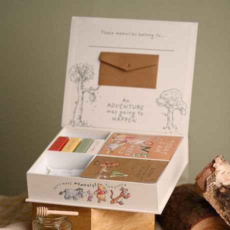 Open white Winnie the Pooh keepsake box showing "These memories belong to..." lid, a kraft envelope, and twelve milestone cards in a divided tray with Pooh and friends illustrations.