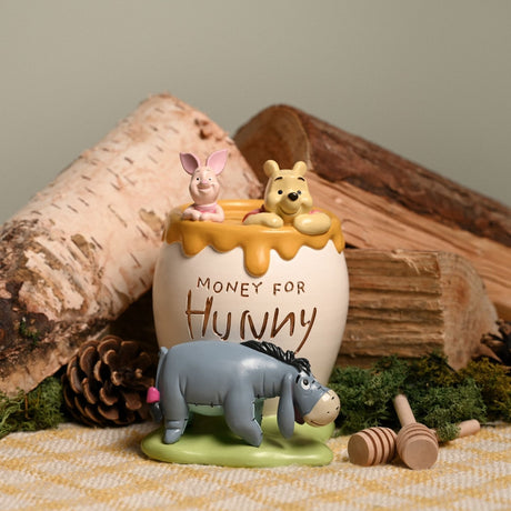 White resin honey pot money box engraved Money for Hunny, with Winnie the Pooh and Piglet peeking over the dripping rim and Eeyore nearby, styled with logs and pinecones.