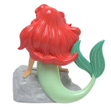 Rear view of the Ariel Mermaid Leaning on Rock resin money box, showing her flowing red hair, green curved tail with fin, and arms resting on a textured grey rock, on a plain white background.