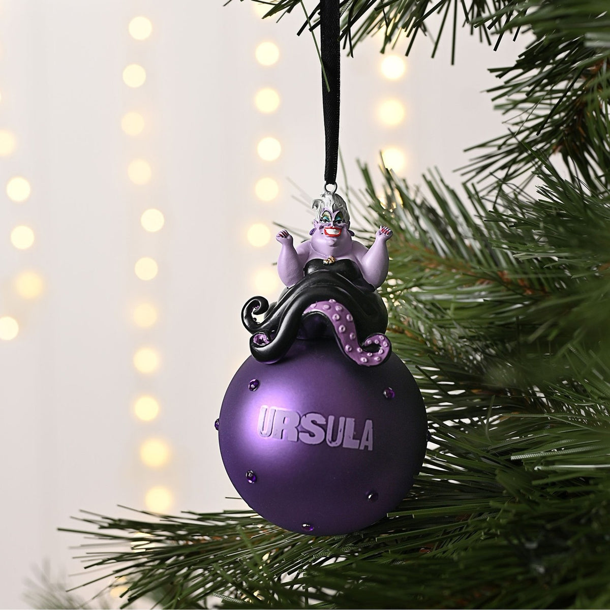Ursula on Glass Bauble Christmas Tree Decoration in purple, with figure perched atop a studded round bauble reading "URSULA", hung from a black ribbon on a Christmas tree.