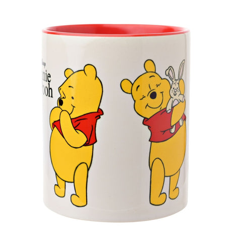White stoneware Winnie the Pooh mug with red interior, showing the bear bashful with paws to chin and cuddling a toy rabbit, on a plain white background.