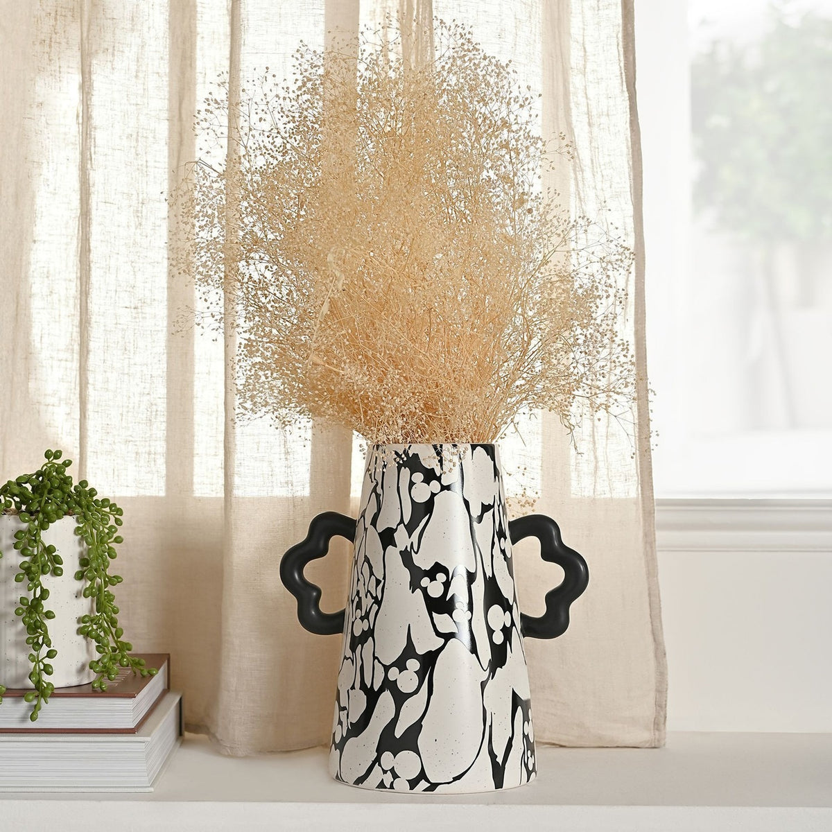 Large ceramic vase with black marbled Mickey Mouse pattern on speckled white, tapered body and wavy black handles, holding dried grasses on a living-room sideboard.