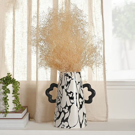 Large ceramic vase with black marbled Mickey Mouse pattern on speckled white, tapered body and wavy black handles, holding dried grasses on a living-room sideboard.