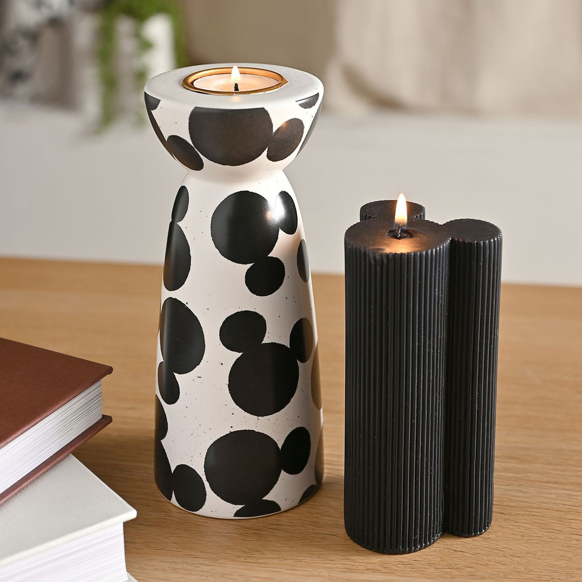 White speckled ceramic tealight holder with scattered black Mickey Mouse shapes, narrow waist, gold insert, lit on wooden table beside books and a black pillar candle.