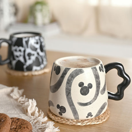 Mickey Mouse Shapes Speckled Stoneware Mug in white with charcoal grey swirls and Mickey head motifs, black curved handle, shown filled on a jute coaster with a matching patterned mug blurred behind.