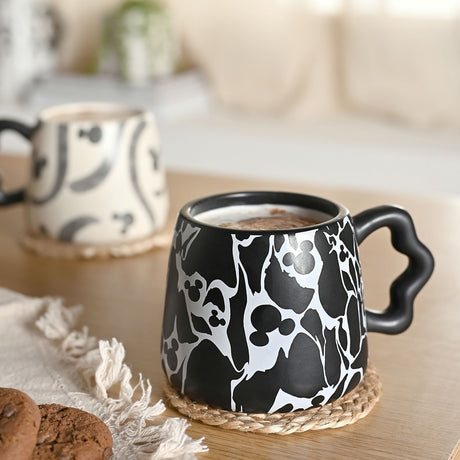 Black stoneware mug with white crackle pattern and hidden Mickey Mouse head silhouettes, wavy handle, shown filled with a hot drink beside a patterned mug on a wooden table.