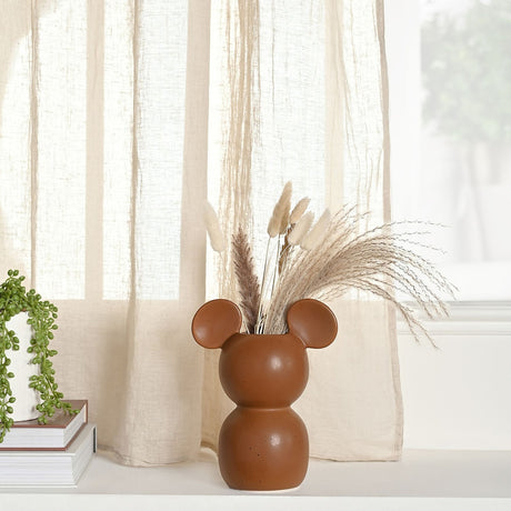 Mickey Mouse shaped ceramic vase in speckled brown glaze with rounded ear openings, holding dried pampas grass on a shelf beside books and sheer curtains.