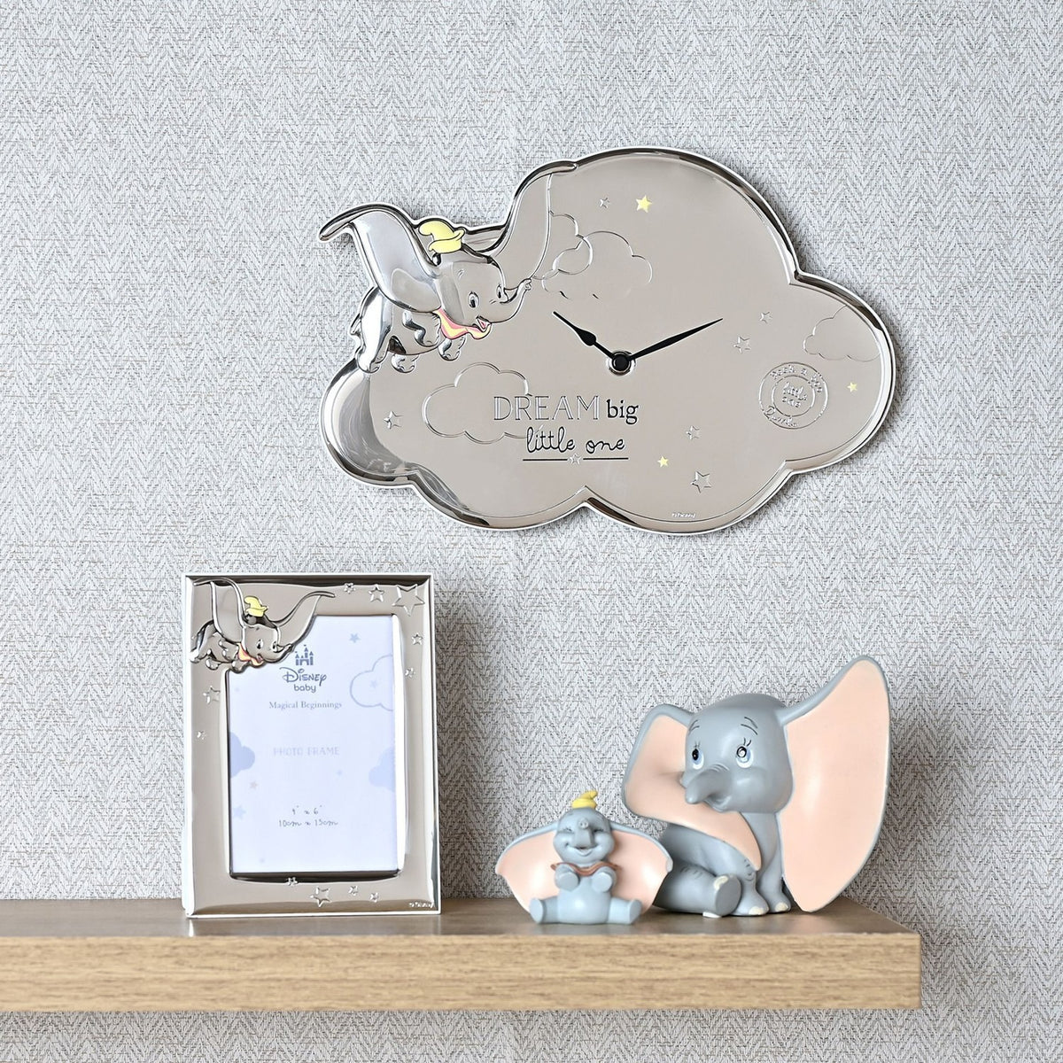 Silver cloud-shaped wall clock featuring Dumbo in relief, embossed stars, and DREAM big little one text, displayed above a shelf with a matching photo frame and Dumbo figurines in a nursery.