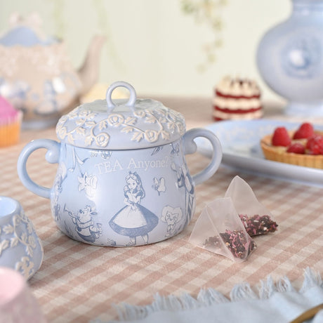 Light blue stoneware tea caddy with an embossed floral lid, twin handles, and sketched Alice in Wonderland scenes, displayed on a table with teabags.