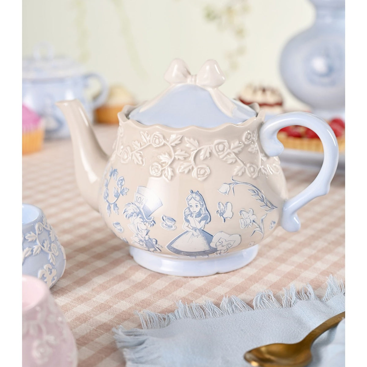 Alice in Wonderland stoneware teapot in beige with light blue Alice and Mad Hatter illustrations, raised rose band, bow-topped lid, and curved handle, shown on a tea table setting.