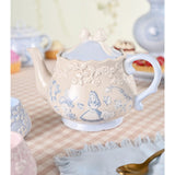 Alice in Wonderland stoneware teapot in beige with light blue Alice and Mad Hatter illustrations, raised rose band, bow-topped lid, and curved handle, shown on a tea table setting.