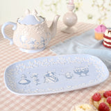 Rectangular blue glazed earthenware serving plate with white floral border and Alice in Wonderland characters in the centre, shown on a tea table beside a teapot and cakes.